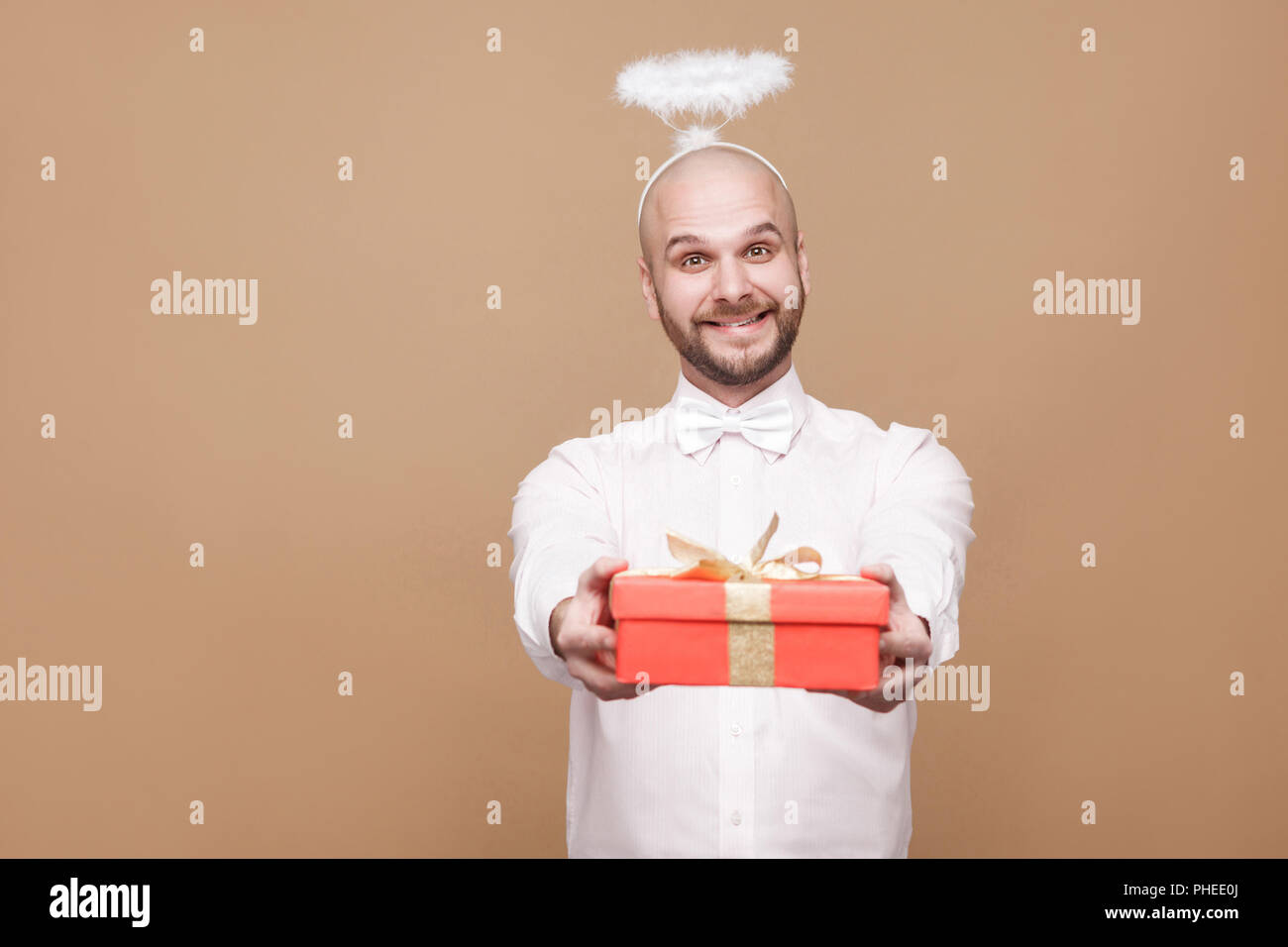 Happy handsome middle aged bald bearded angel in shirt and white halo on head standing and giving red gift and looking at camera with toothy smile. st Stock Photo