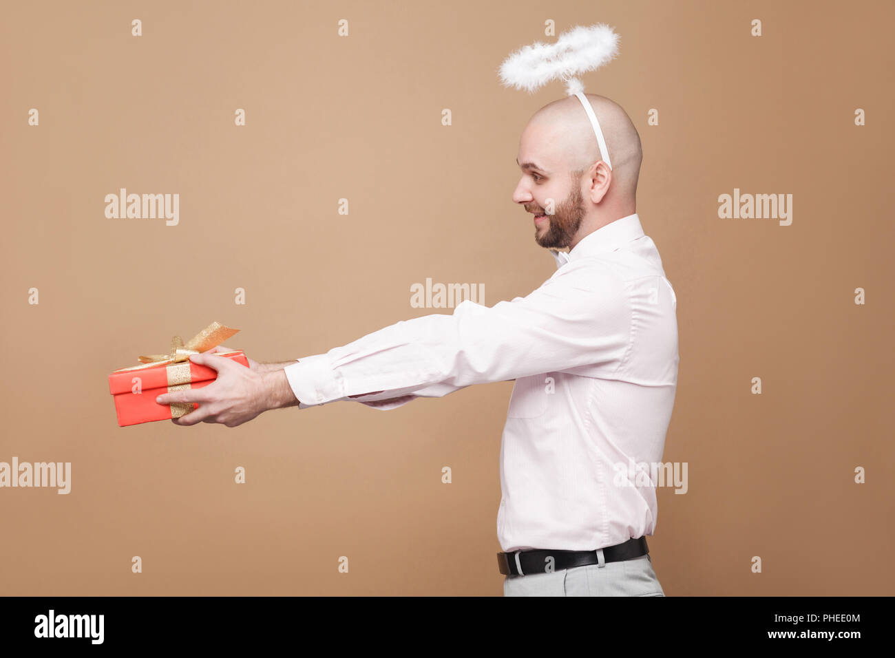 profile side view of happy handsome middle aged bald bearded angel in shirt and white halo on head standing and giving red gift with toothy smile. stu Stock Photo