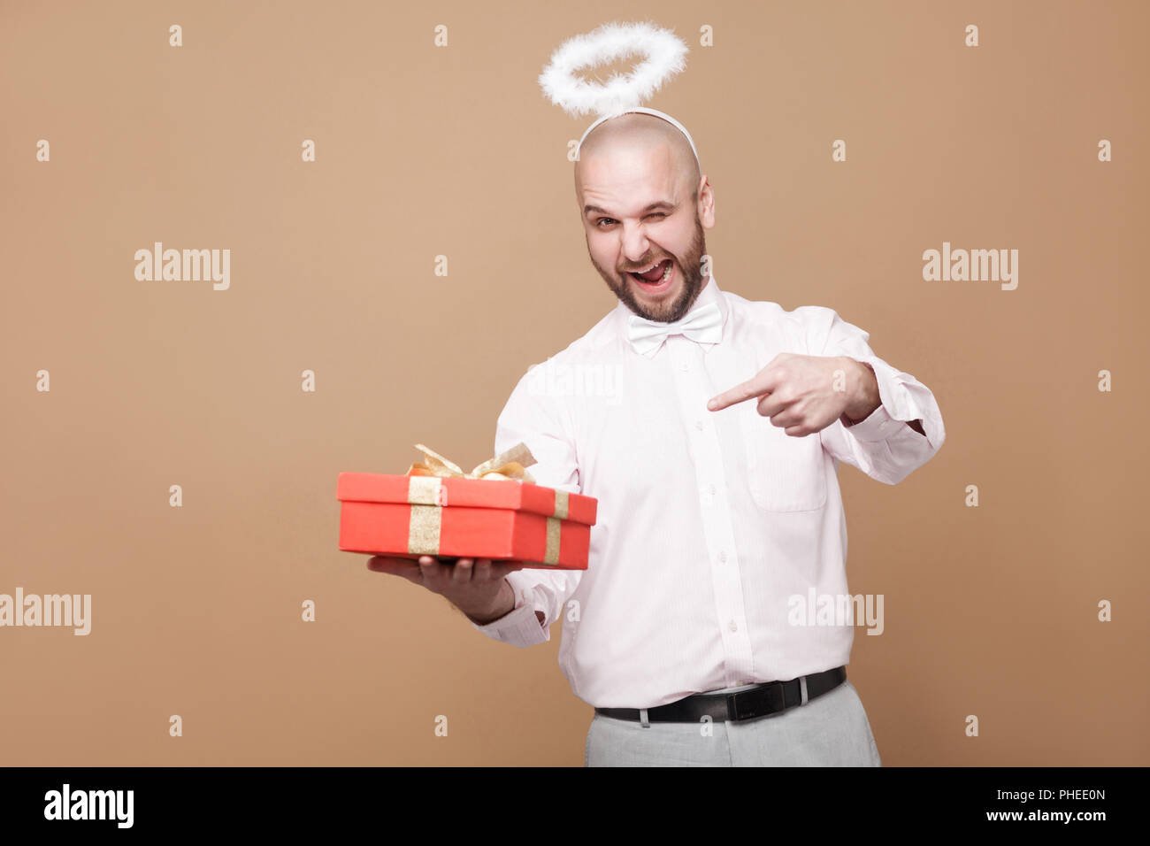 This is for you. Happy funny middle aged bald bearded angel in shirt and white halo on head pointing at red gift and winking with smile. indoor studio Stock Photo