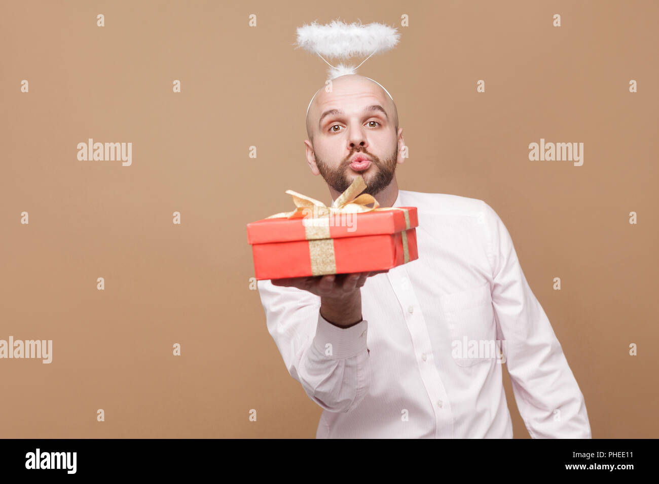 Happy handsome middle aged bald bearded angel in shirt and white halo on head standing holding red gift, looking at camera and sending kiss. indoor st Stock Photo