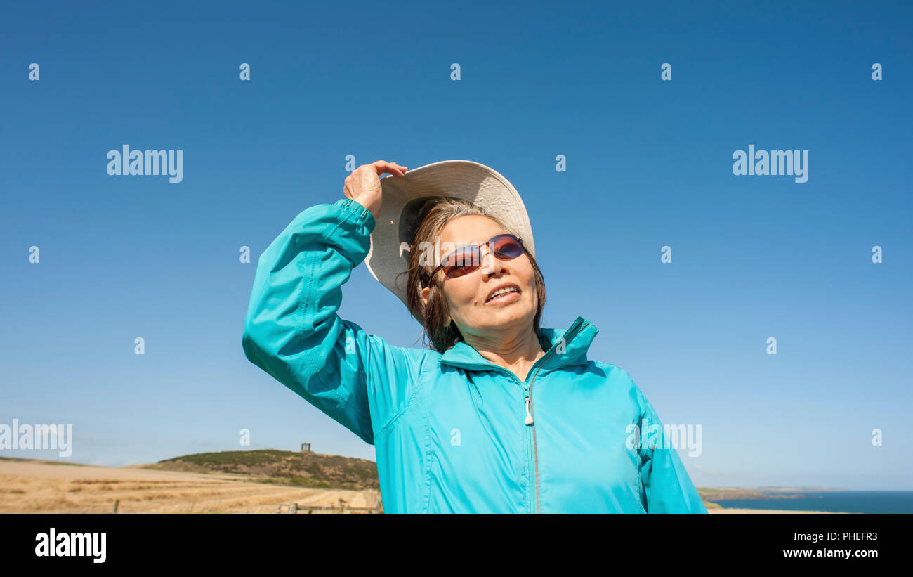 Attractive mature Asian lady wears blue anorak, canvas hat, sunglasses. Looks up to blue sky with  facial expression for interpretation. Stock Photo