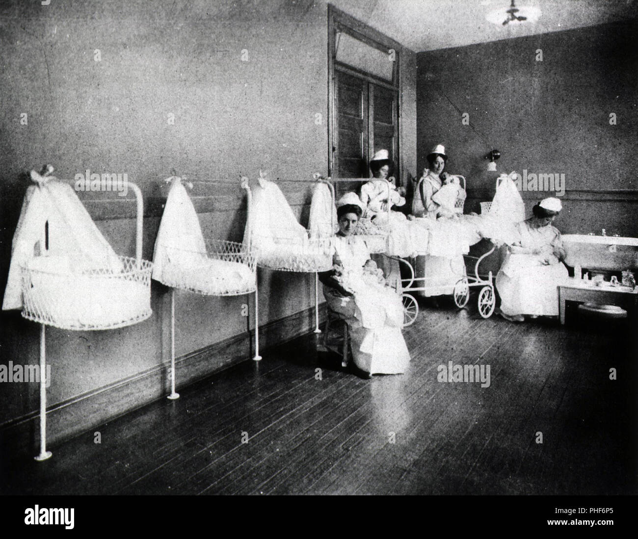 Four nurses hold infants in a large room with baby baskets mounted along one wall. Stock Photo