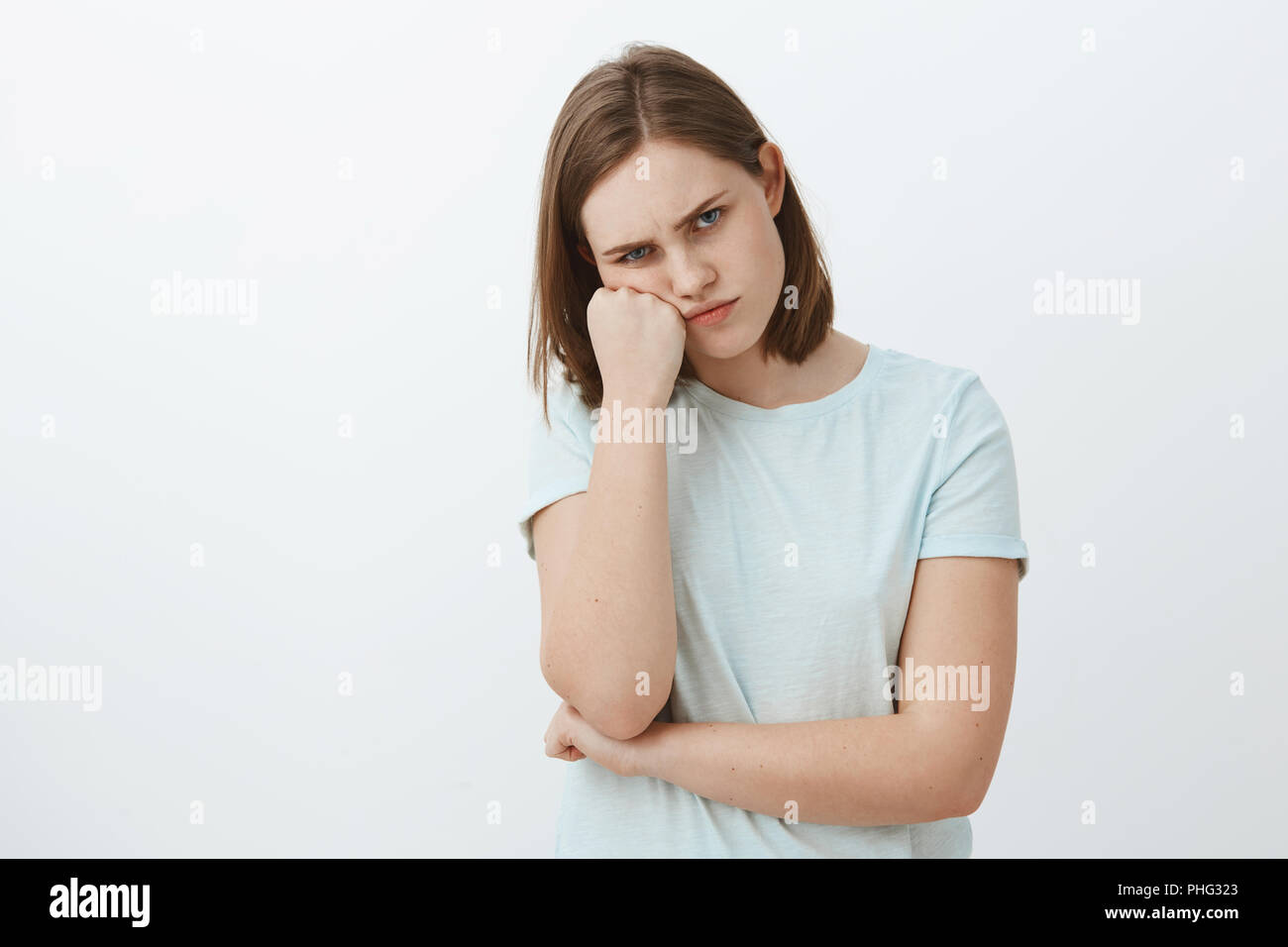 Studio shot of displeased offended and gloomy cute female brunette in t-shirt leaning face on fist frowning and pursing lips feeling envy or jealous seing sibling received awesome gift from parents Stock Photo