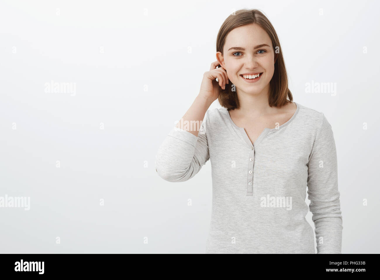 Cute emotive teenage girl in casual blouse flicking hair behind flapped ear and smiling broadly feeling shy and joyful being invited to play with interesting company over white background Stock Photo