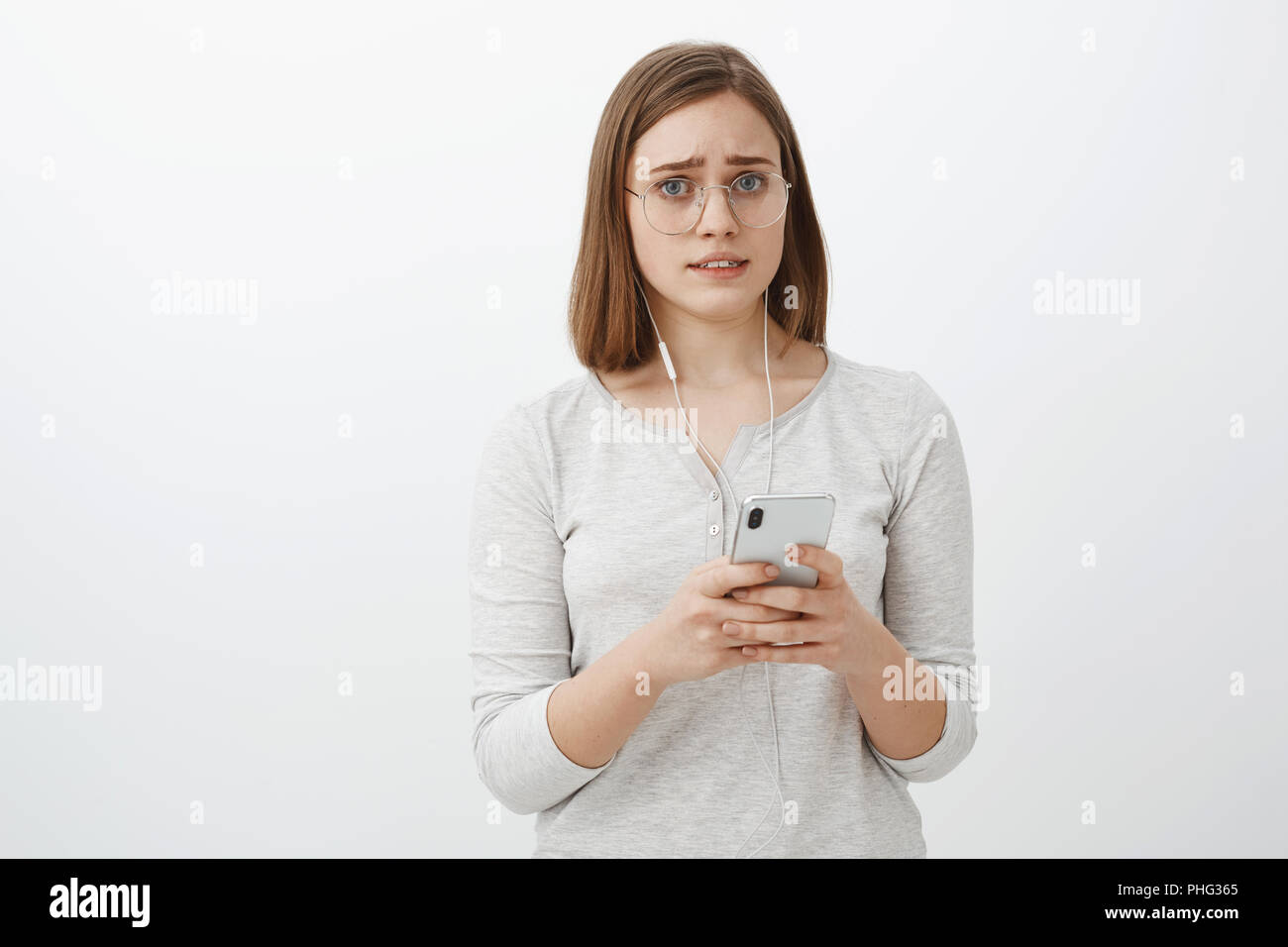 Girl feeling worried one earphone broke down. Portrait of displeased and concerned upset cute woman in glasses with short brown hair frowning and making sad face holding smartphone wearing headphones Stock Photo