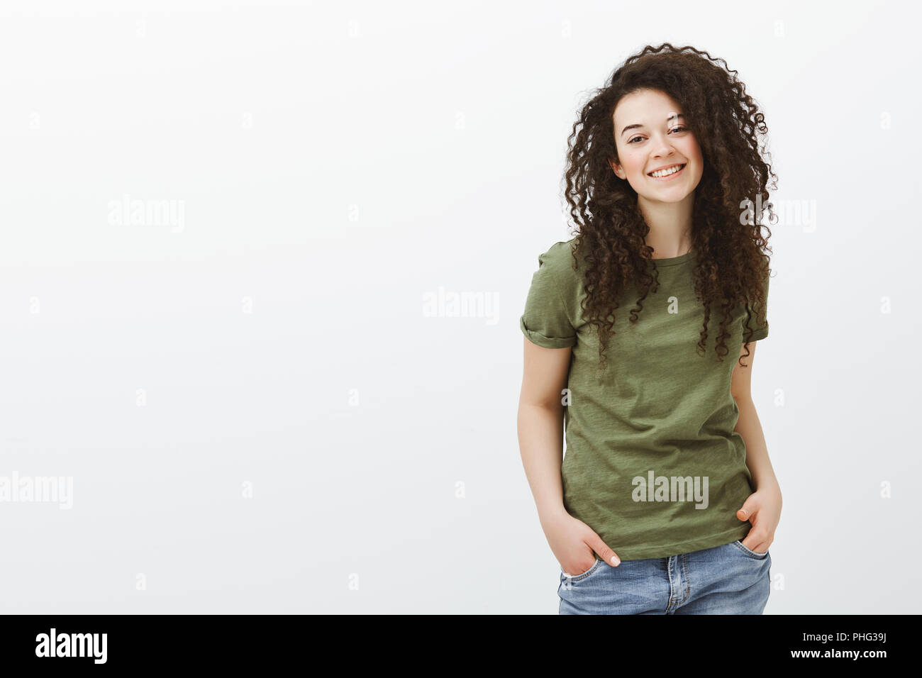 Portrait of joyful good-looking adult woman with curly hair, hanging out over gray background, holding hands in pockets and smiling with happy friendly attitude, talking casually with friends Stock Photo