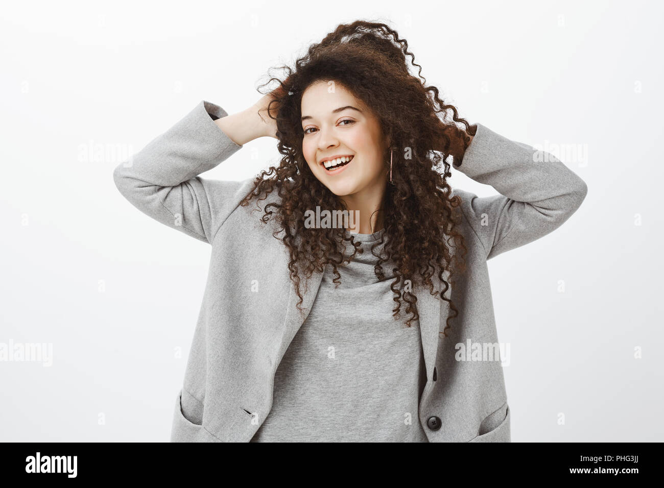 Girl feeling releived and carefree after visiting spa. Joyfull bright european female with curly hair in grey coat, touching curls and smiling broadly, expressing awesome and pleased attitude Stock Photo