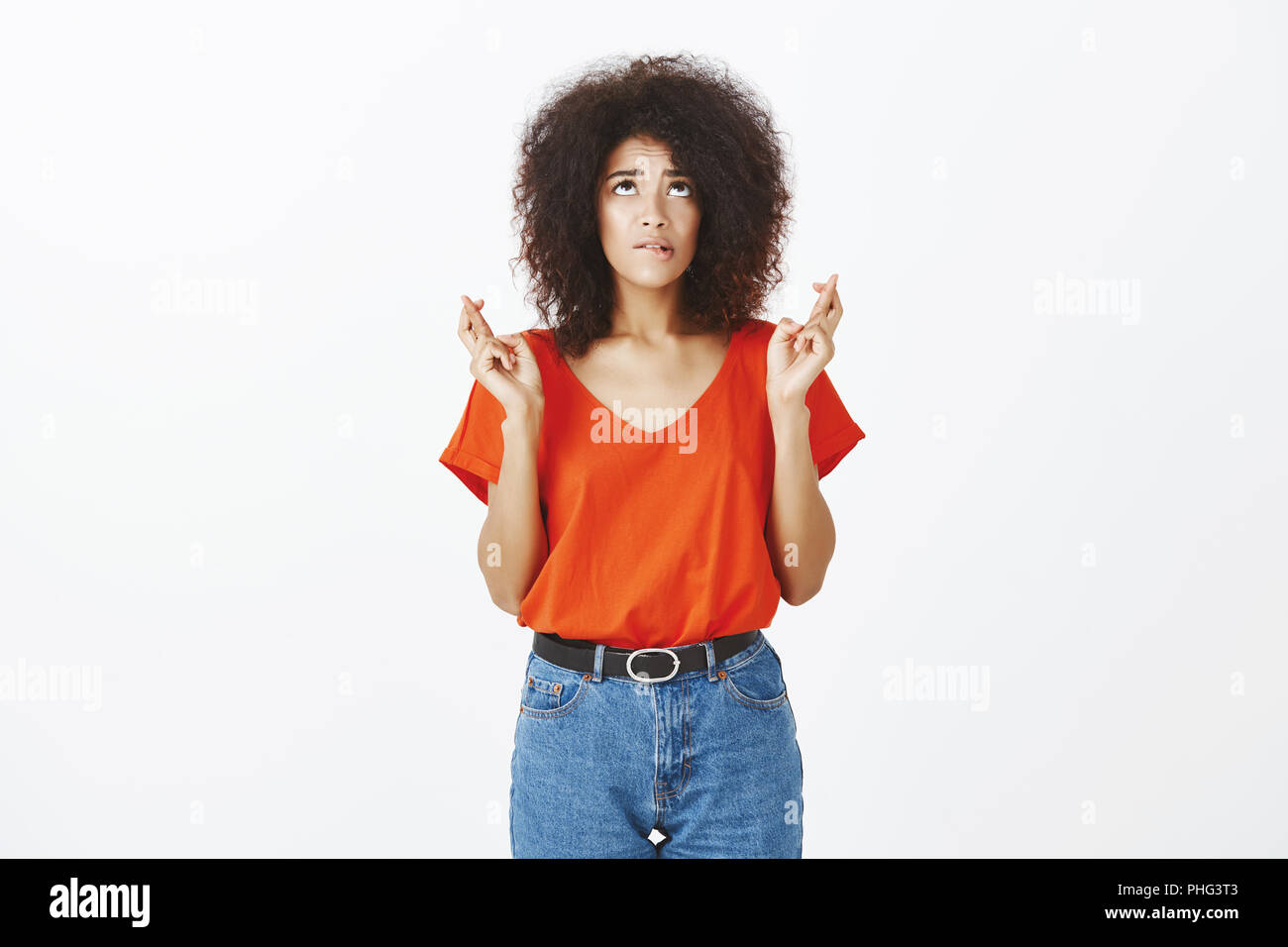 Indoor shot of intense worried female student with afro hairstyle, looking up and biting lip, standing with crossed fingers, begging or praying, making wish and wanting something badly Stock Photo