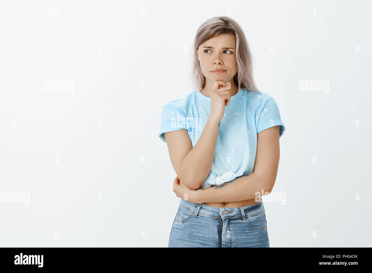 Girl feeling not sure about received suggestion. Doubtful intense charming woman with fair hair, frowning and pursing lips while holding hand on chin and looking aside, thinking, making choice Stock Photo