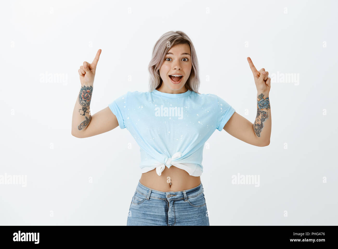 Portrait of excited joyful charming woman with tattooed arms, pointing up with raised index fingers and smiling broadly while discussing fantastic news, standing impressed and happy over gray wall Stock Photo