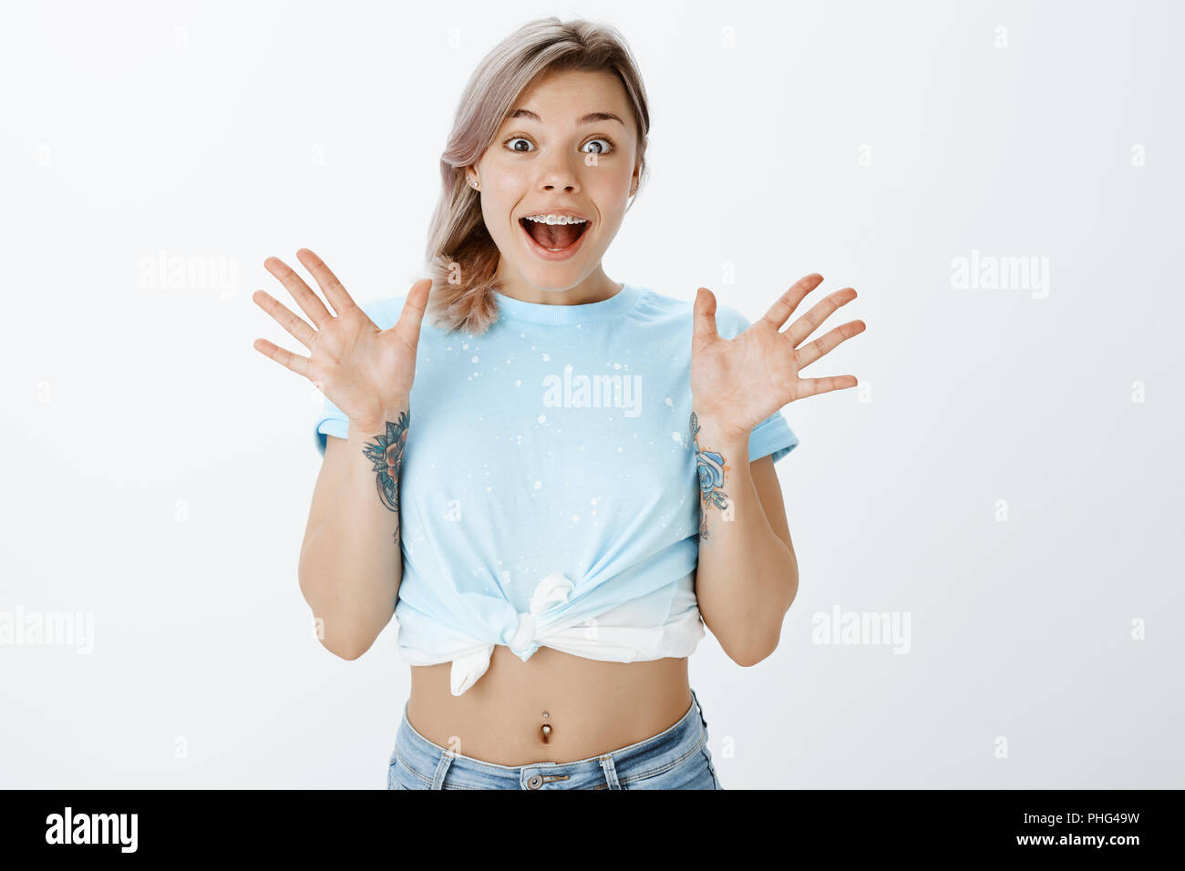 I am so lucky, cannot believe. Portrait of surprised and excited good-looking woman in cropped t-shirt with tattooed arms and pierced belly, raising palms from happiness and smiling broadly Stock Photo