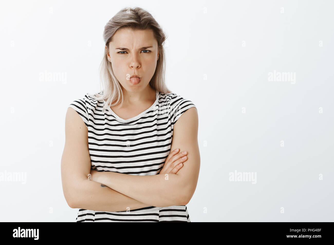 I do not want talk with you. Portrait of disobedient angry young woman with fair hair, sulking and frowning, sticking out tongue and holding hands crossed, being offended or insulted, arguing Stock Photo