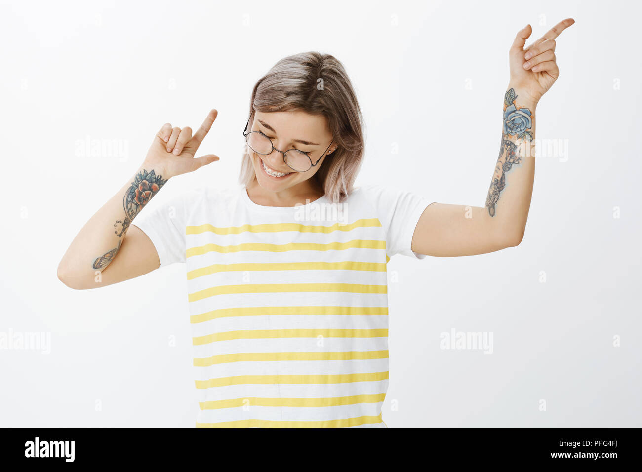 Dance and enjoy life. Portrait of positive happy young girlfriend in striped tellow t-shirt, waving and gesturing with tattooed arms, smiling joyfully while gazing downwards, being in great mood Stock Photo