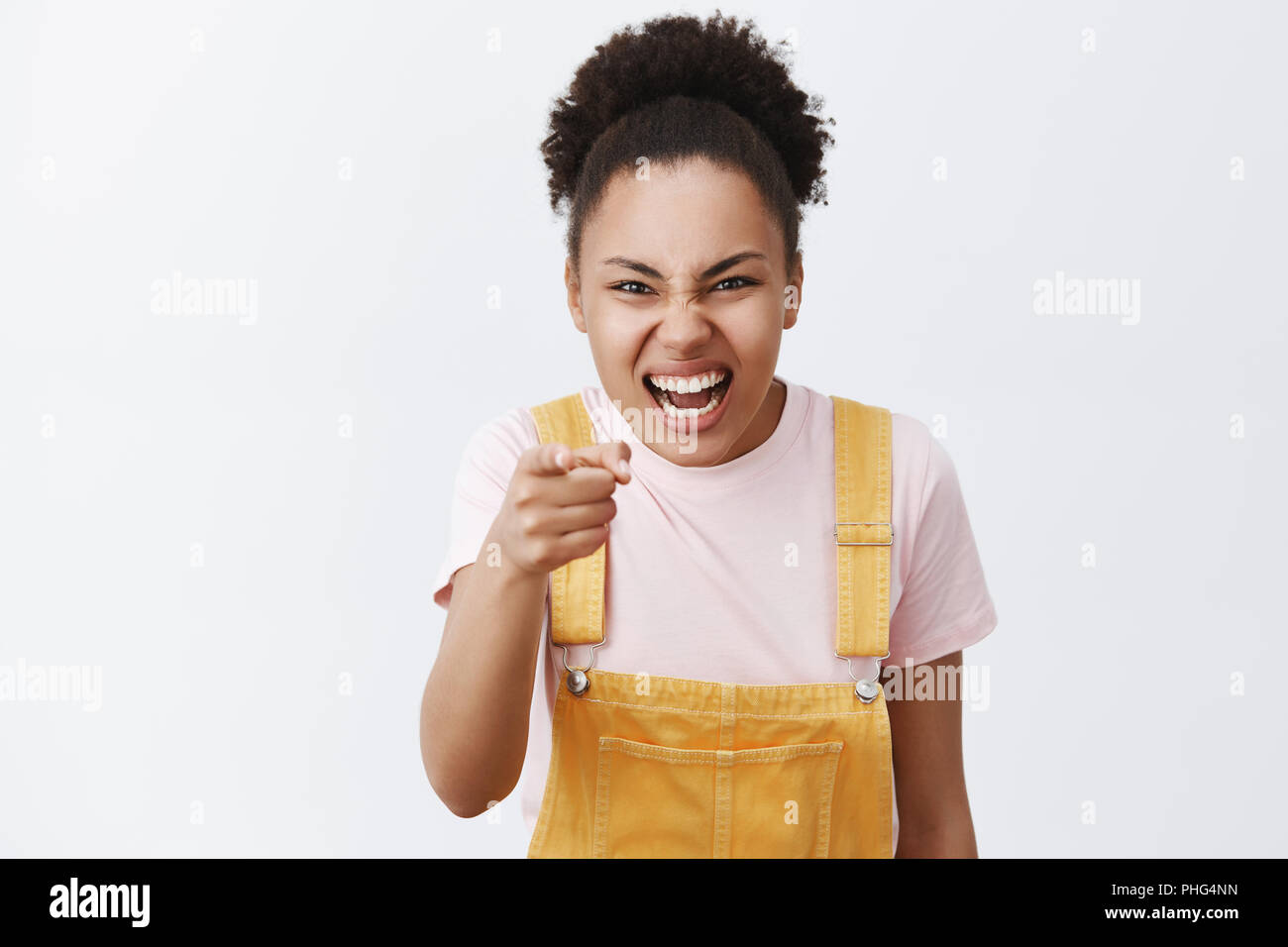 Ha-ha you are ugly. Portrait of rude impolite female bully with dark skin in yellow overalls, expressing scorn and disdain, pointing at camera and laughing over someone over gray background Stock Photo