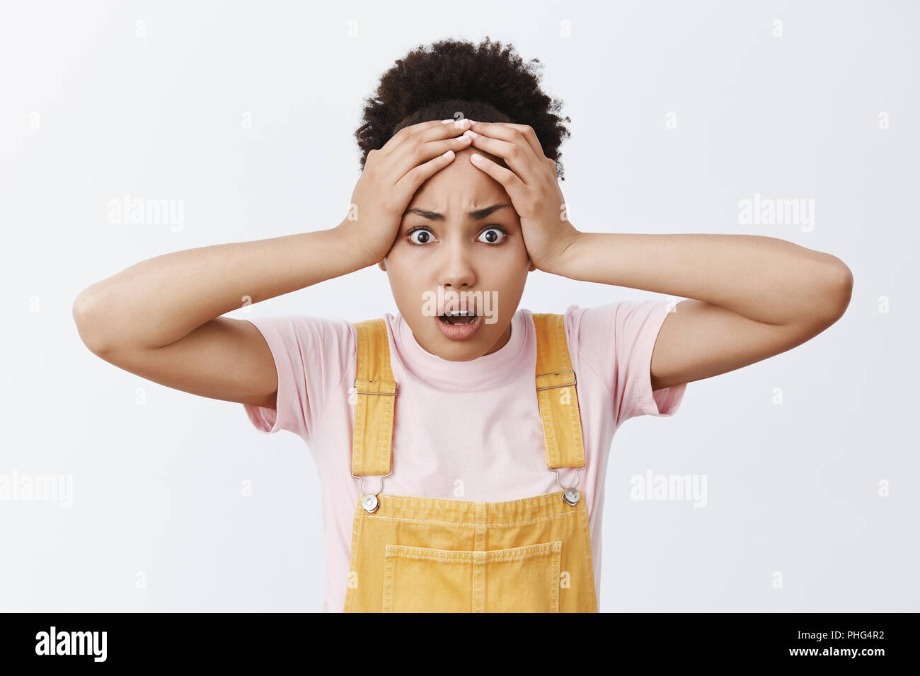 Portrait of shocked and terrified troubled cute woman with dark skin in trendy yellow overalls, holding hands on forehead and staring with fright and anxiety at camera, worrying over grey wall Stock Photo