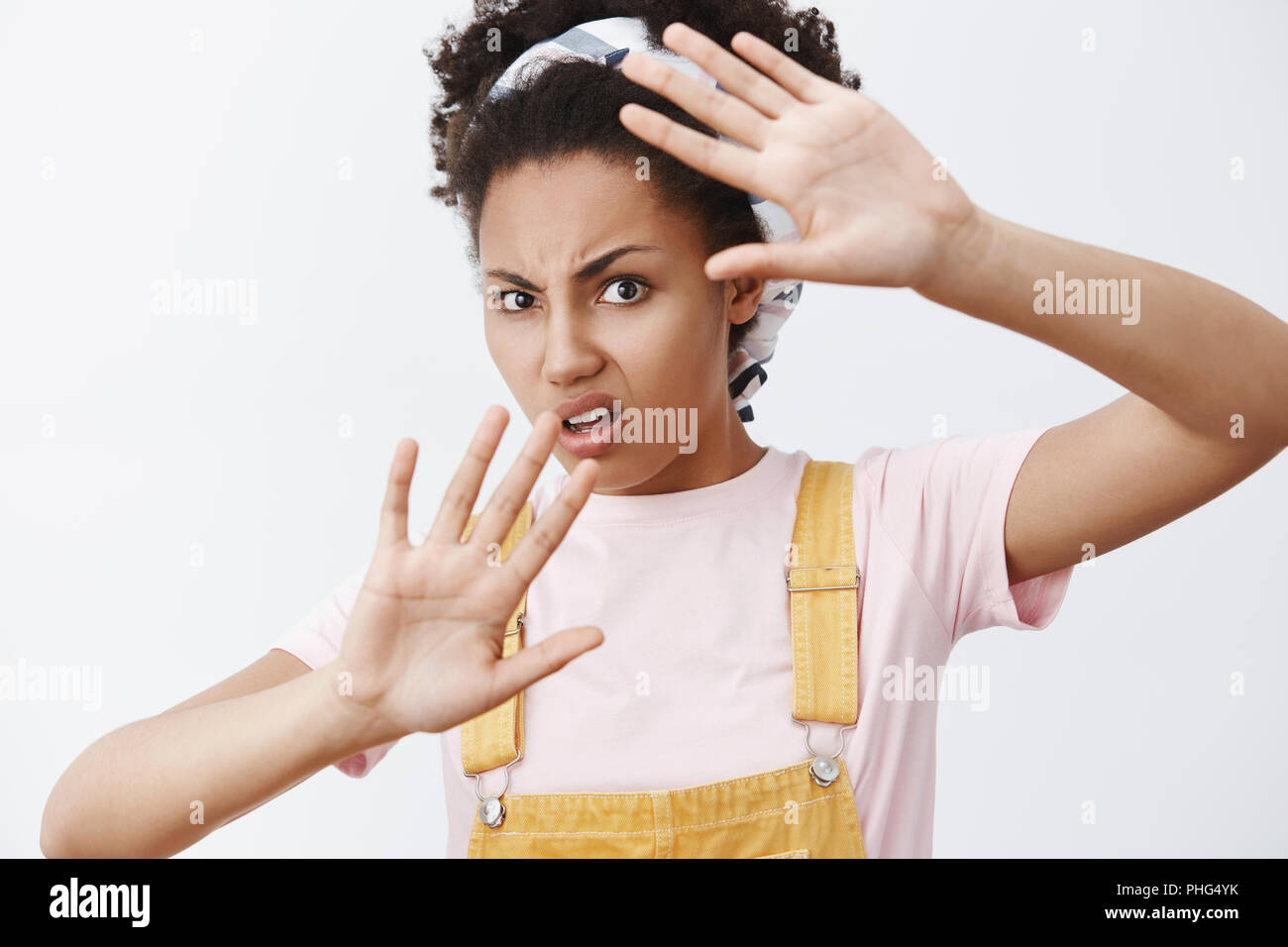 Stop waving it around my face. Portrait of displeased and bothered annoyed African American modern female in yellow overalls and headband, covering from flash of camera with raised palms, frowning Stock Photo