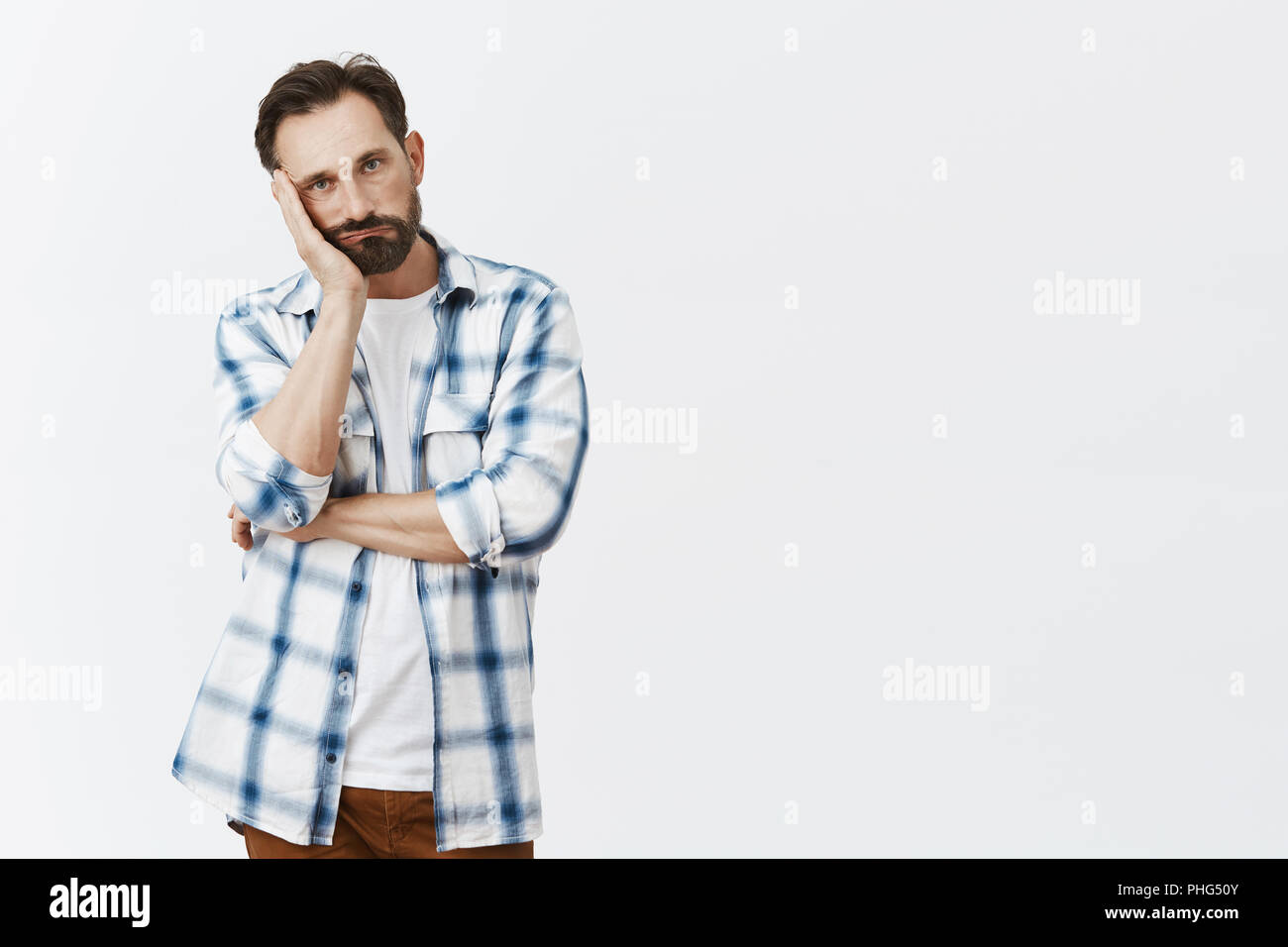 Adult man starting losing temper, leaning face on palm, pursing lips and staring with annoyance and boredom, being displeased and bothered with uninteresting talk, standing indifferent over grey wall Stock Photo