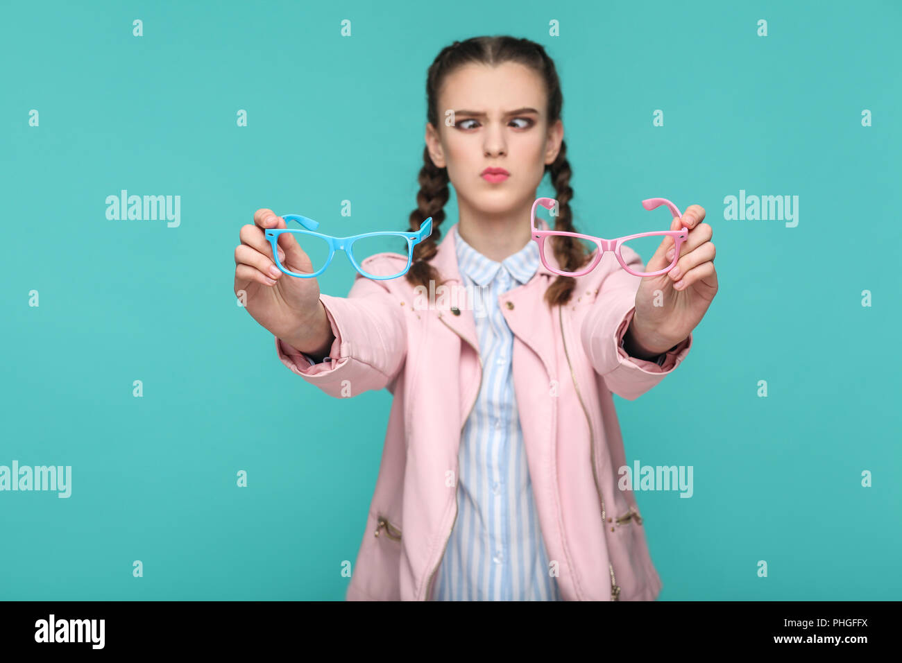 doubtful funny girl in casual or hipster style, pigtail hairstyle, standing, holding blue and pink glasses and looking at camera with crossed eye, Ind Stock Photo