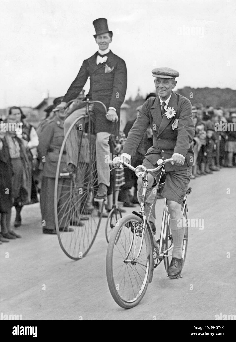 Penny farthing bicycle. A man is riding a penny-farthing bicycle besides a man on a regular bicycle. 1930s Stock Photo