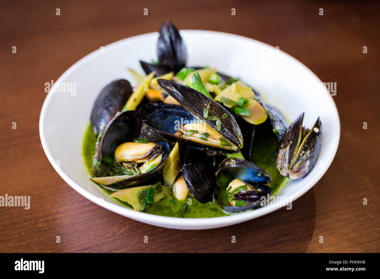 Mussels in shells in a white bowl with a green sauce on a dining table. Stock Photo