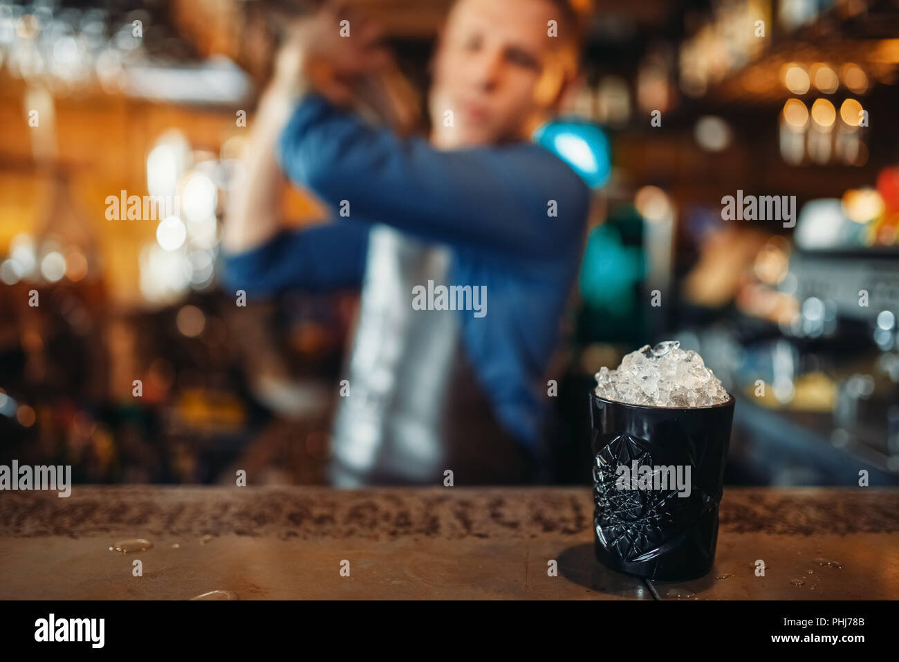 Black glass full of ice stands on bar counter, male barman with shaker on background. Barkeeper occupation, bartender job Stock Photo