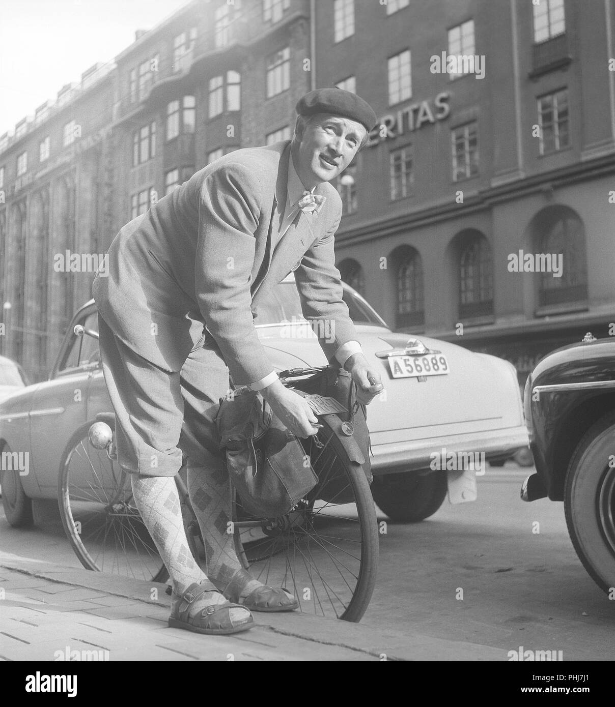 1950s man with his bicycle. A man in has parked his bicycle and balancing it on the kerb. He puts a chain around the back wheel, unabling it from being stolen. Notice the registration sign on the bicycle that was a mandatory item at this time.  Sweden 1954. Photo Kristoffersson ref BP57-5 Stock Photo