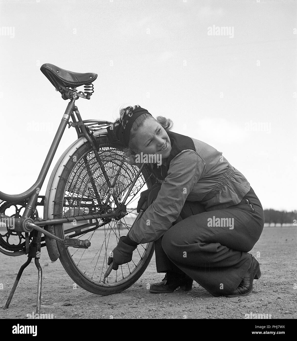1940s couple with a bicycle. A young woman is pumping air into the back tire of her bicycle. She is wearing the typical sports clothing of the 1940s with long trousers and a short jacket. Notice the protection net mounted over the back wheel to prevent hanging clothes from getting caught in the wheel. Sweden 1942.  Photo KristofferssonA89-1 Stock Photo