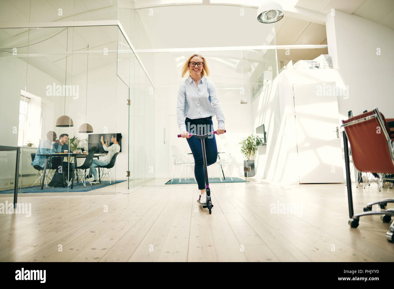 Smiling young businesswoman riding a scooter around the floor of a ...