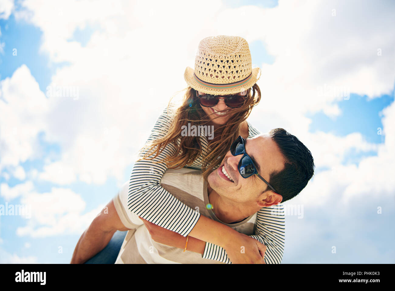 Handsome young man with his loving wife or girlfriend having fun relaxing on summer vacation piggy back riding against a sunny cloudy blue sky Stock Photo