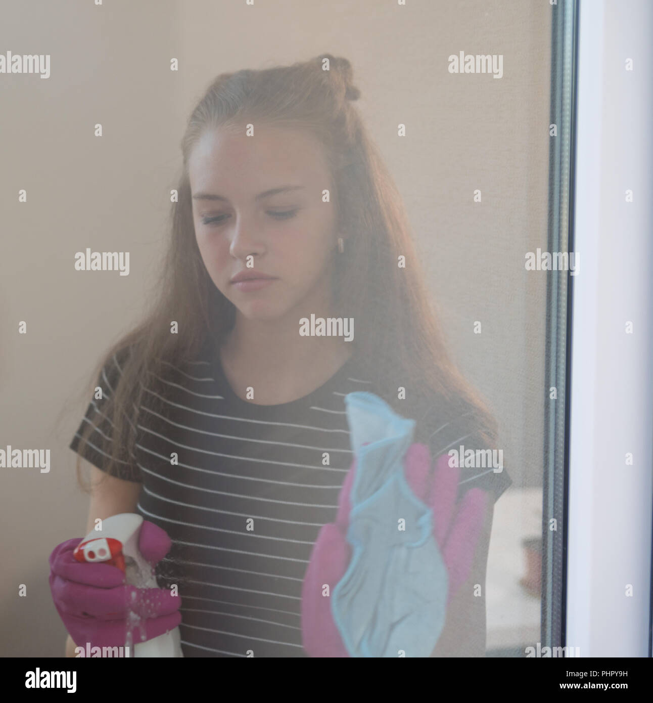 Sulky young woman cleaning a window pane with a spray bottle and cloth viewed through the glass