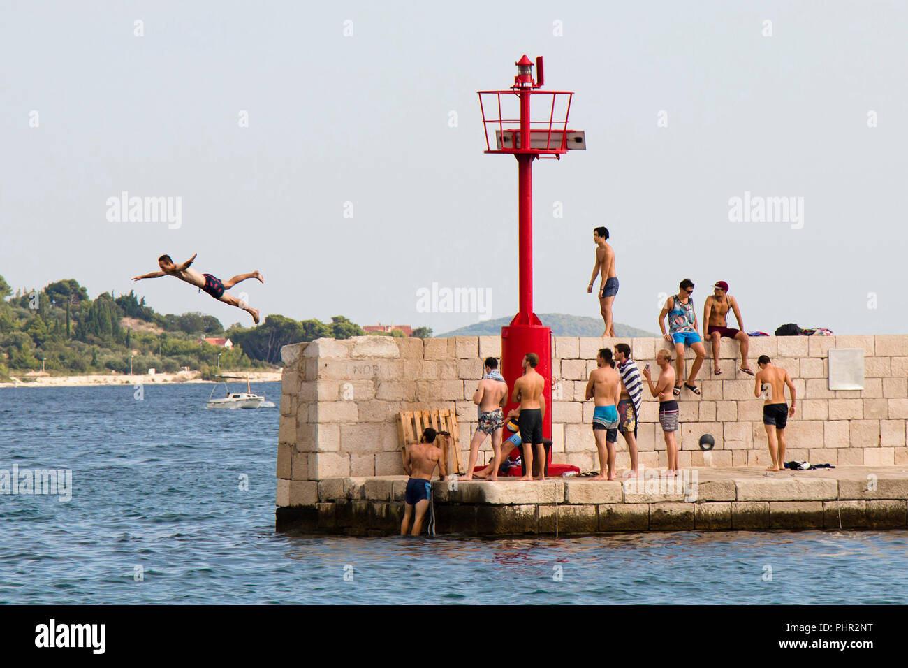 Vodice, Croatia - August 2, 2018: Young men in swimsuits standing on the pier and watching a man jumping in the sea in summer season Stock Photo