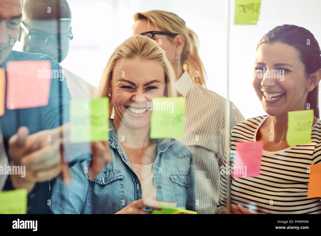 Diverse group of businesspeople laughing while standing in an office brainstorming together with sticky notes on a glass wall Stock Photo