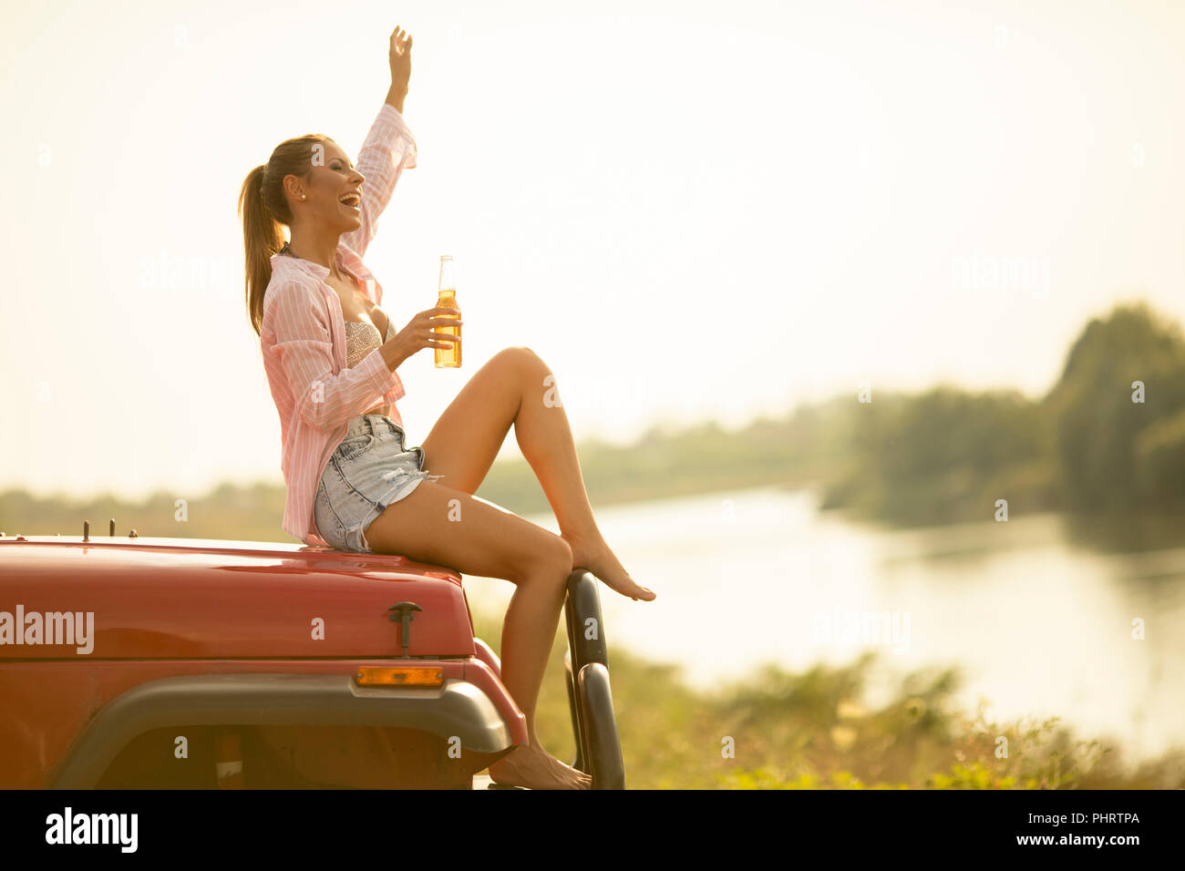 Young woman sits on a car hood,  drinks refreshment and enjoys it Stock Photo