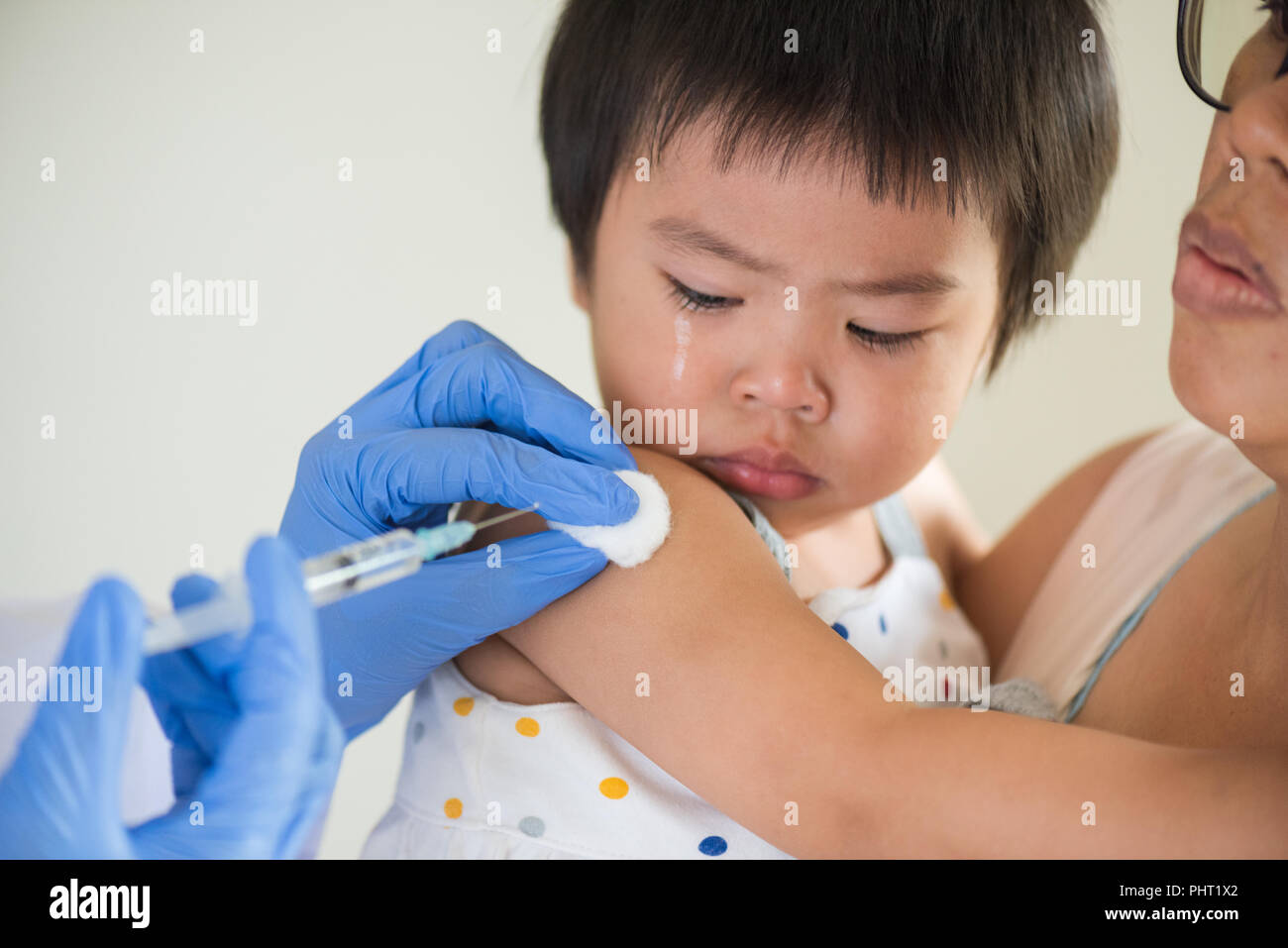 Doctor giving an injection vaccine to a girl. Little girl crying with her mother on background. Stock Photo