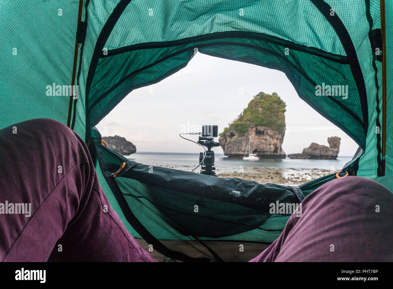 Lifestyle image with legs in purple jeans on foreground of POV view from a tent on the beach of the bay with rocks. Action Camera on tripod. Traveling filmmaker lifestyle. Stock Photo