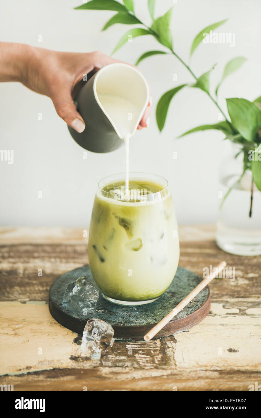 https://c8.alamy.com/comp/PHTBD7/iced-matcha-latte-drink-in-glass-with-coconut-milk-pouring-from-pitcher-by-womans-hand-white-wall-and-plant-branches-at-background-summer-refreshin-PHTBD7.jpg