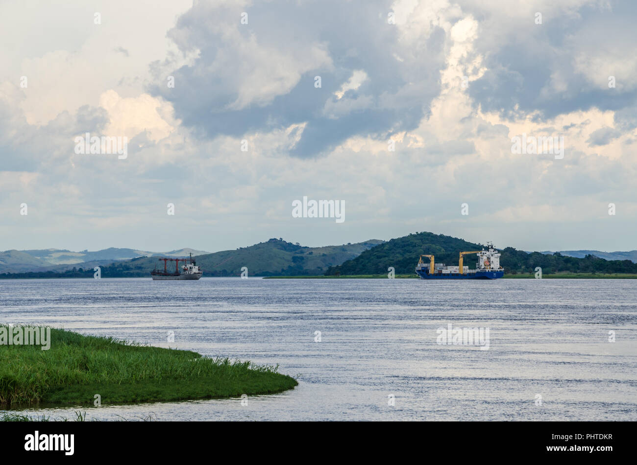 Container cargo ships on mighty Congo river with dramatic cloudy sky and lush green grass in foreground, Democratic Republic of Congo, Africa. Stock Photo