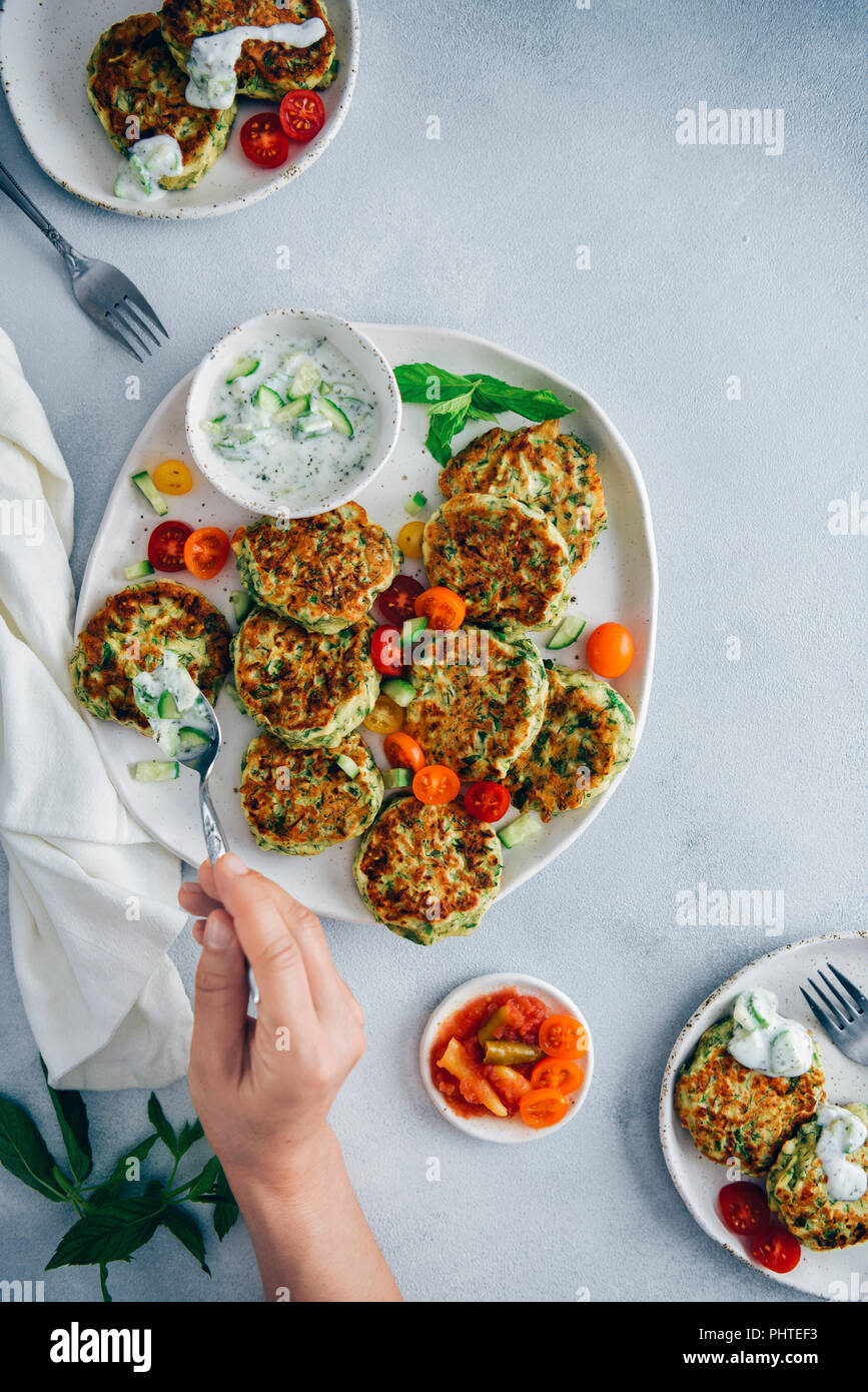 Hand spreading cucumber yogurt dip on zucchini fritters served on a white ceramic plate photographed from top view. Stock Photo