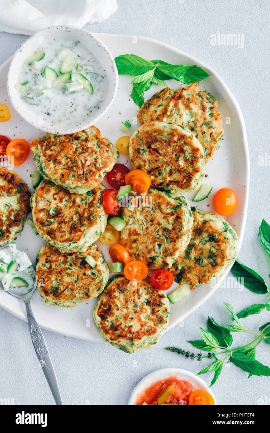 Zucchini fritters on a ceramic plate served with grape tomatoes and yogurt dip on the side photographed from top view. Stock Photo