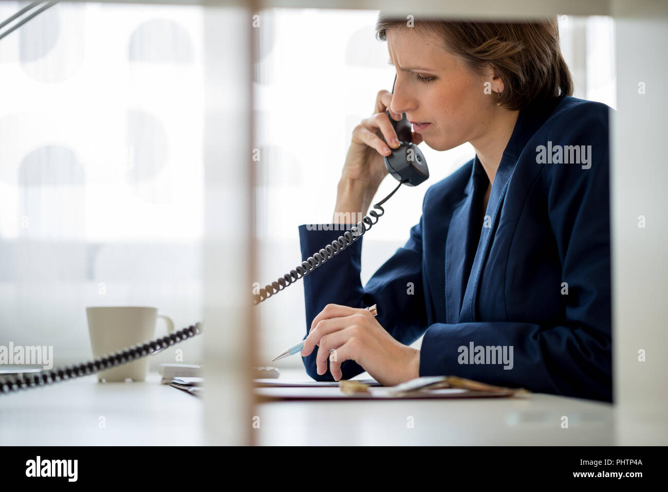 Businesswoman or manageress talking on a landline phone with a worried expression while looking at documents. View through an interior glass partition Stock Photo