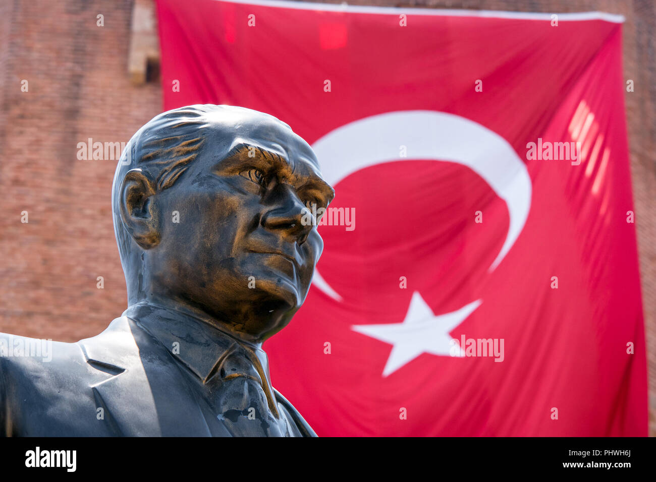 A close up of a proud bronze statue of Mustafa Kemal Atatürk in front of a large red Turkish flag hanging from a building. Stock Photo