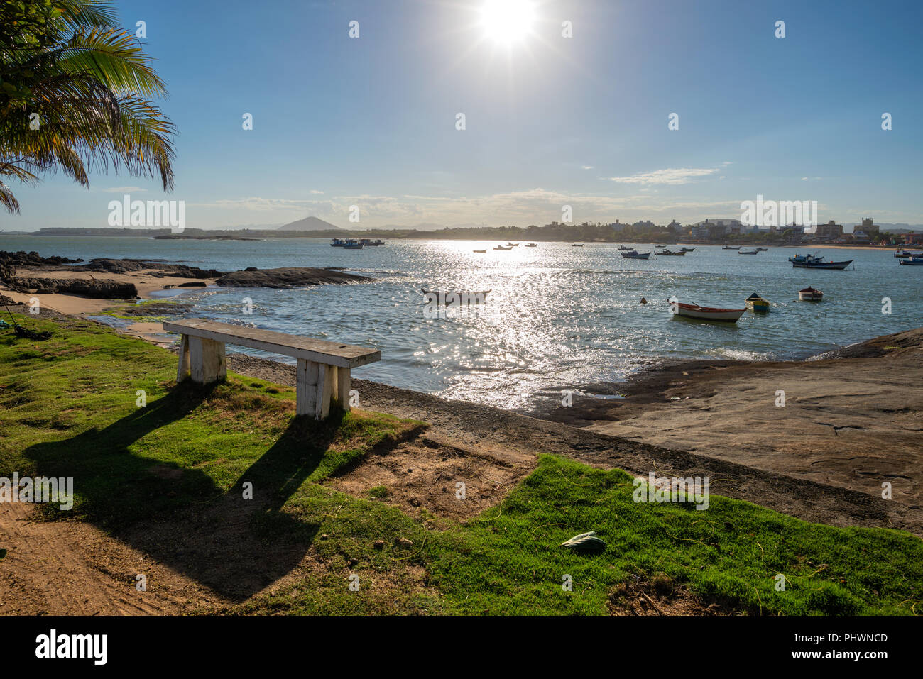 Rustic bench in front of the sea with boats and the sun in the background. Meaipe beach, Guarapari, Espirito Santo, Brazil. Stock Photo