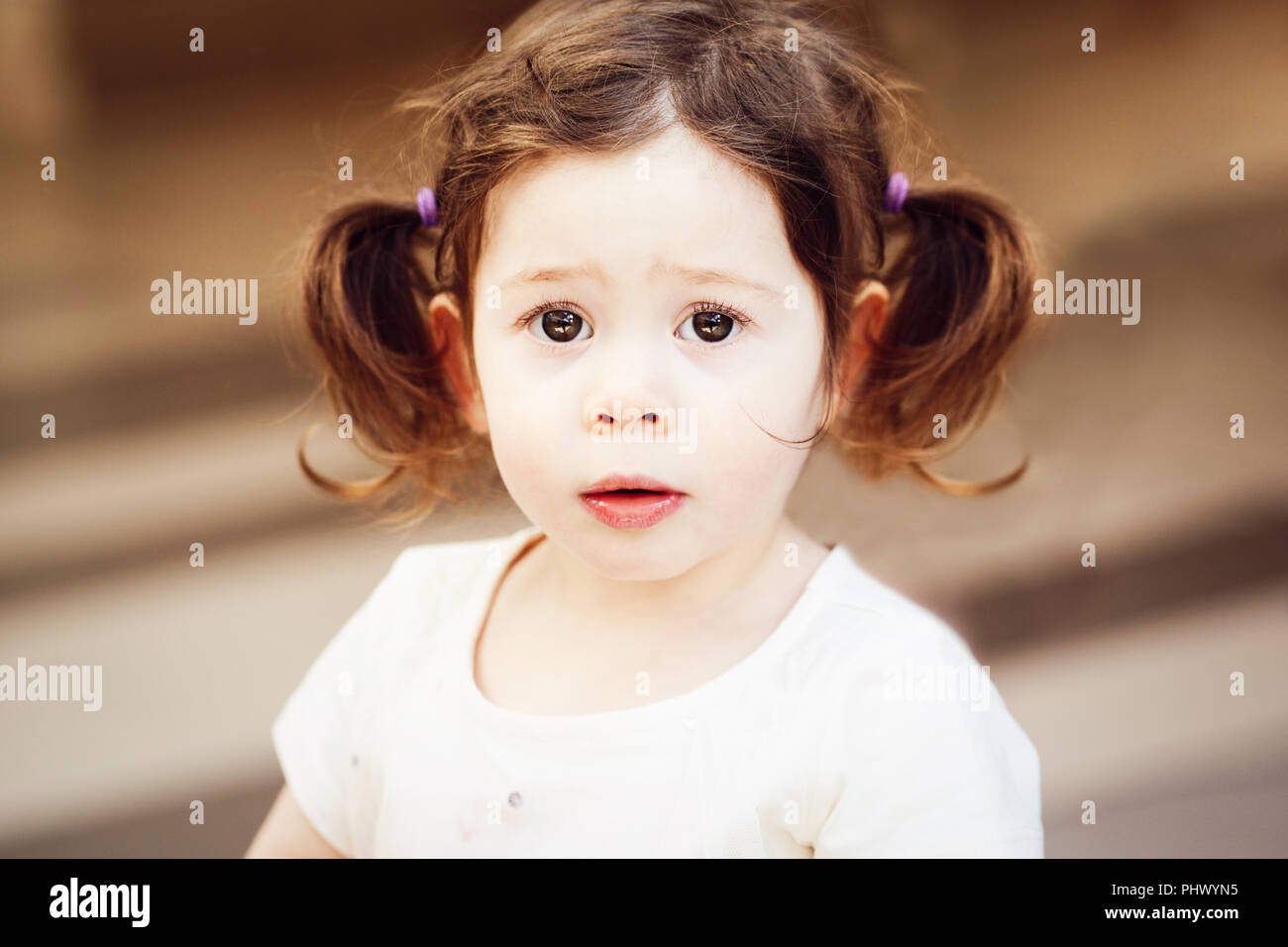 Closeup portrait of cute adorable sad upset white Caucasian toddler girl child with dark brown eyes and curly pig-tails hair in white light dress tshi Stock Photo