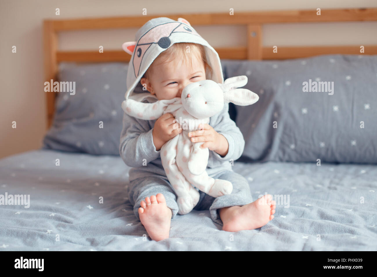 Portrait of cute adorable Caucasian blonde smiling baby girl with blue eyes in grey pajama with fox cat animal hood sitting on bed in bedroom and hold Stock Photo