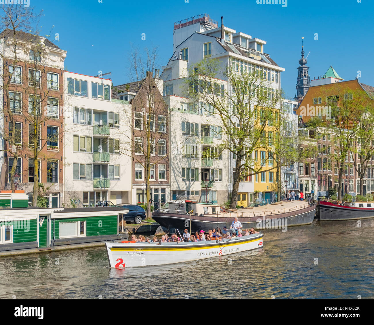 A canal tour boat glides past colourful buildings and houseboats in a canal in Amsterdam Holland. Stock Photo