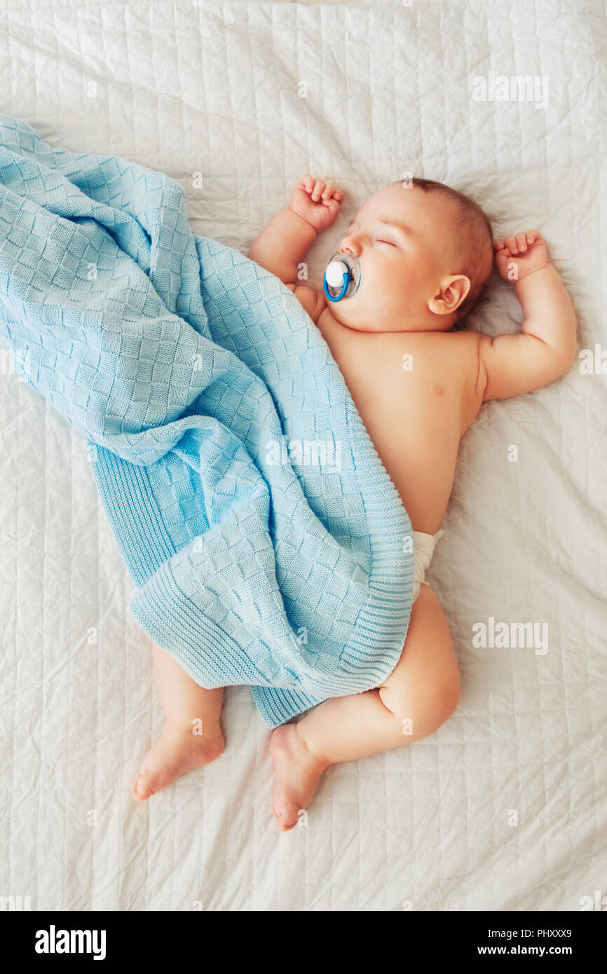 Portrait of a cute adorable white Caucasian baby newborn in diaper, sleeping dreaming with pacifier soother in mouth, lying on bed, covered with blue  Stock Photo