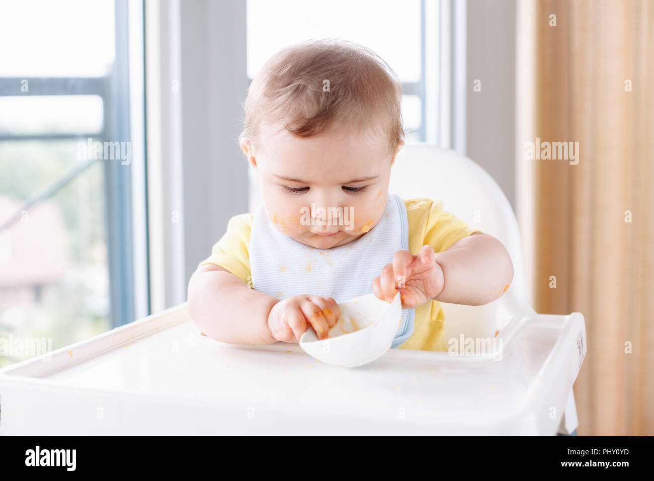 Portrait of cute adorable Caucasian child boy with dirty messy face sitting in high chair eating apple puree with fingers. Everyday home childhood lif Stock Photo