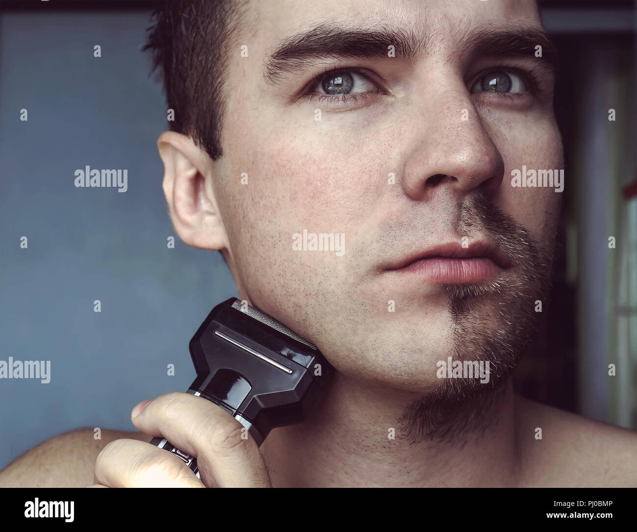 Young bearded man during grooming of beard using trimmer. Half face with a beard half shaved. Stock Photo