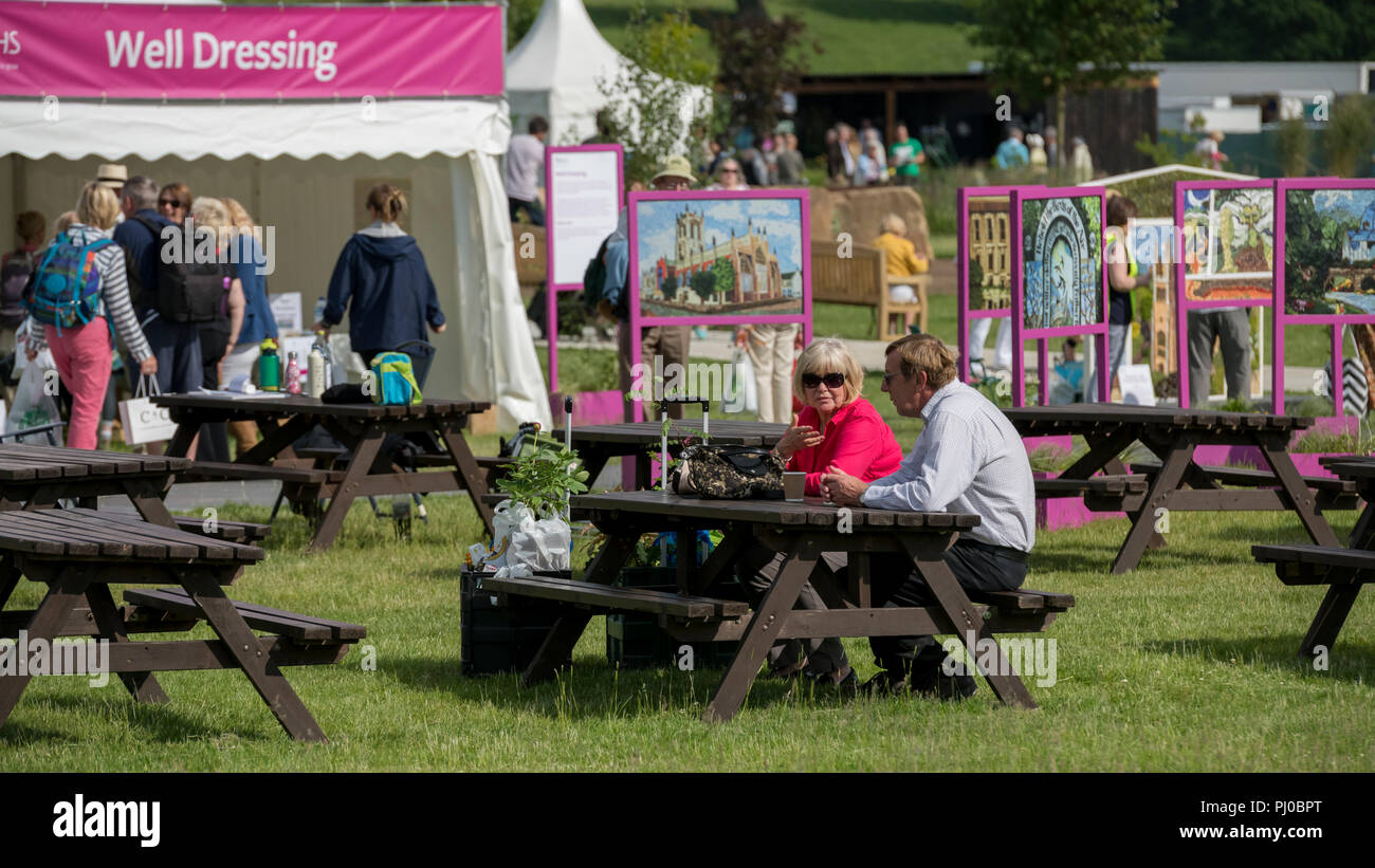 Visitors look round as couple sitting at picnic table have drink & chat by well dressing display - RHS Chatsworth Flower Show, Derbyshire, England, UK Stock Photo