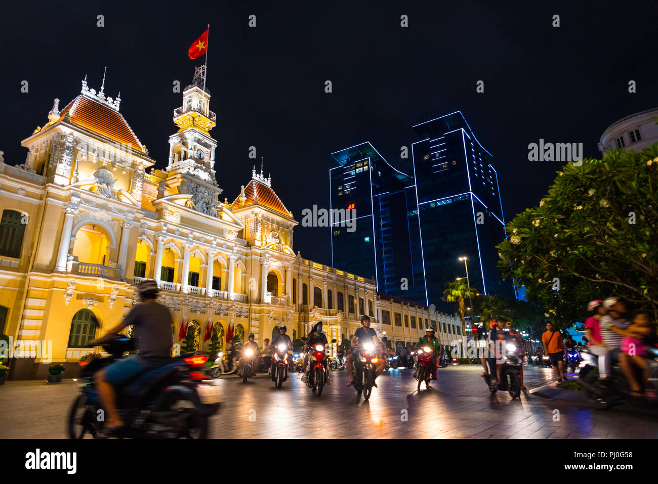 Ho Chi Minh City, Vietnam - May 1, 2018: illuminated Saigon City Hall, Vincom Center towers, colorful street traffic & tropical tree against night sky Stock Photo